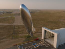 Large shiny airship suspended beside a modern hangar on a desert airfield at sunset