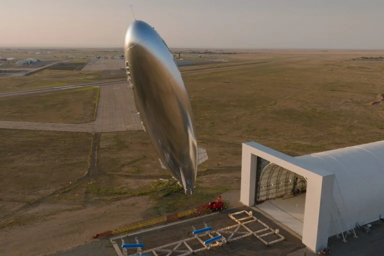 Large shiny airship suspended beside a modern hangar on a desert airfield at sunset