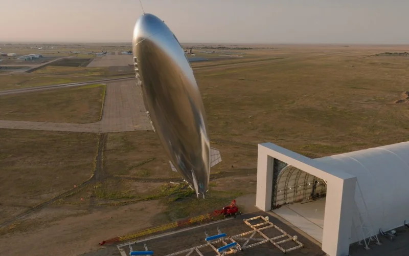 Large shiny airship suspended beside a modern hangar on a desert airfield at sunset