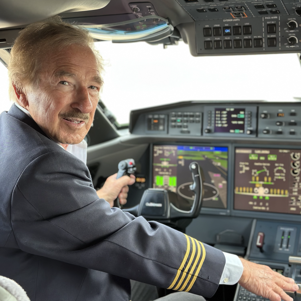 Steven F Udvar Hazy Pilot in a dark uniform with gold stripes on the sleeve sits in the cockpit hand on the control yoke with digital flight displays and overhead panels visible ahead