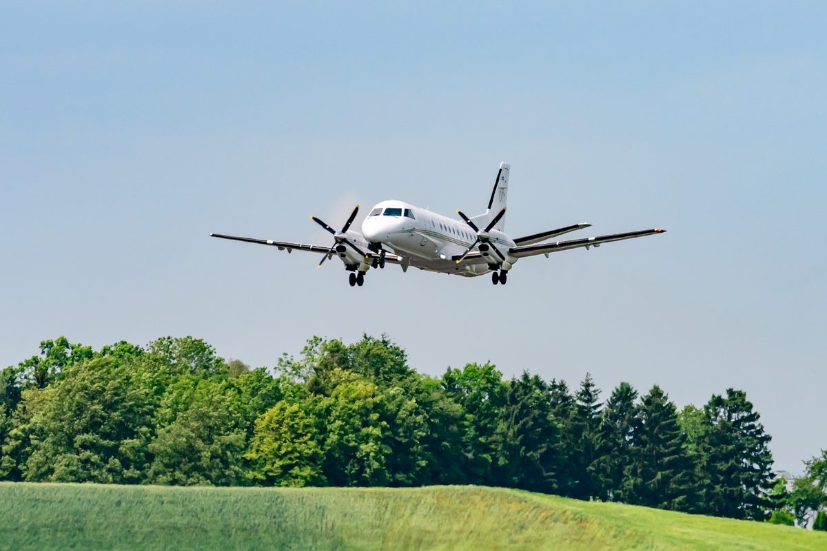 HoerschingAustria21May2024SwedishAirForceSvenskaFlygvapnet White twin engine turboprop airplane flying low over a green field with trees and a clear blue sky behind it approaching for landing