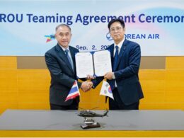 Two businessmen in dark suits shake hands while showing signed agreement documents at a ceremony Thai and South Korean flags on the table with a model helicopter centerpiece