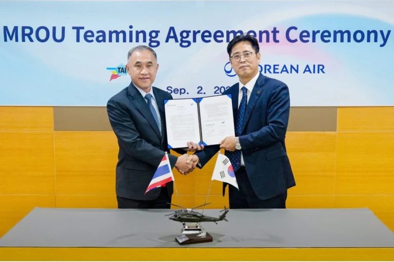 Two businessmen in dark suits shake hands while showing signed agreement documents at a ceremony Thai and South Korean flags on the table with a model helicopter centerpiece