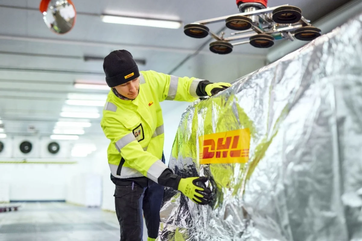 DHL to build healthcare logistics hub at Changi Airport A worker in a high visibility yellow jacket and black beanie smooths reflective insulation foil over a large metallic surface in a warehouse like facility gloves on