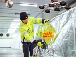A worker in a high visibility yellow jacket and black beanie smooths reflective insulation foil over a large metallic surface in a warehouse like facility gloves on
