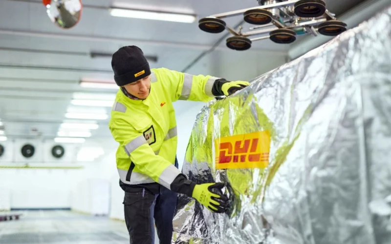 A worker in a high visibility yellow jacket and black beanie smooths reflective insulation foil over a large metallic surface in a warehouse like facility gloves on