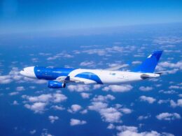 Blue and white commercial airplane cruising above a sea of clouds in a clear blue sky seen from the side of the fuselage and wing