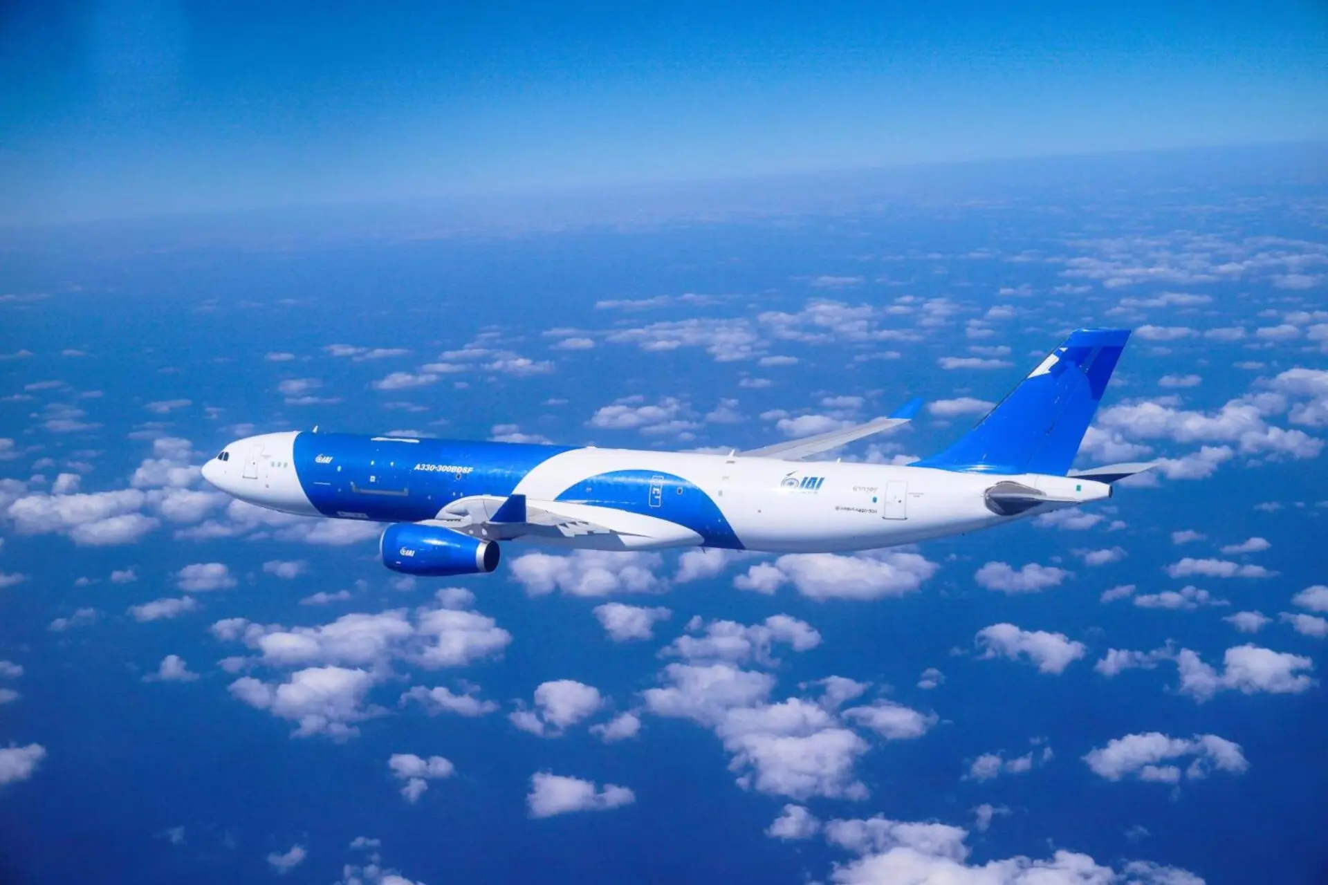 Blue and white commercial airplane cruising above a sea of clouds in a clear blue sky seen from the side of the fuselage and wing