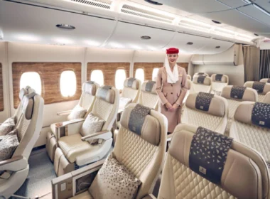 Flight attendant in beige uniform standing in a spacious passenger cabin with beige quilted seats and wood-patterned window panels.