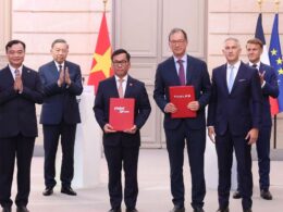 Six men in suits stand on a stage with flags in the background two hold red folders labeled vietjetaircom and THALES