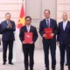 Six men in suits stand on a stage with flags in the background two hold red folders labeled vietjetaircom and THALES