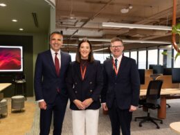 Three professionally dressed colleagues pose for a group photo in a modern office lobby