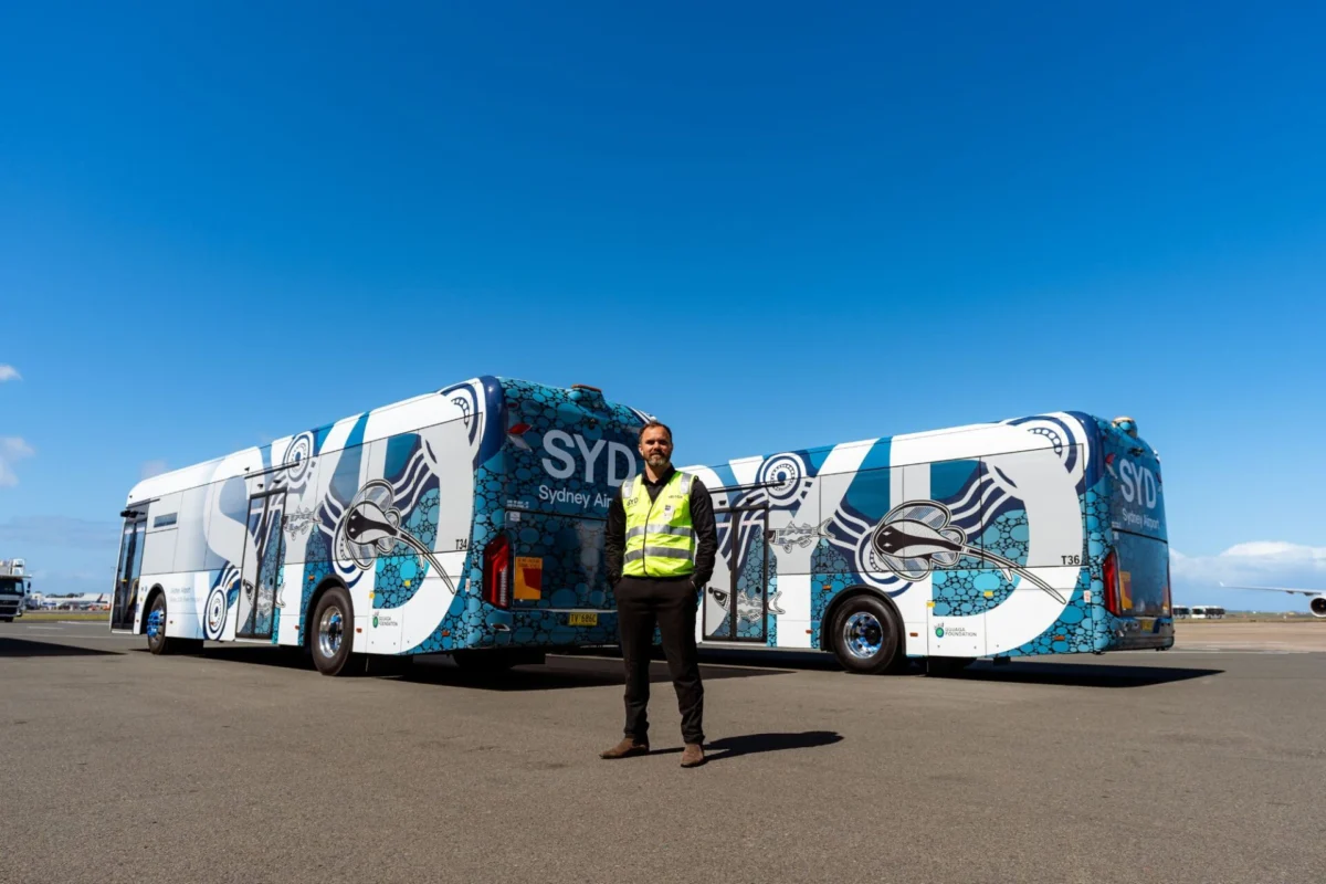Sydney Airport launches electric bus fleet in net zero push Man in a high visibility vest stands in front of a pair of blue and white patterned buses on an airport tarmac under a clear sky