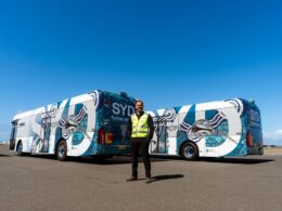 Man in a high visibility vest stands in front of a pair of blue and white patterned buses on an airport tarmac under a clear sky