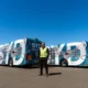 Man in a high visibility vest stands in front of a pair of blue and white patterned buses on an airport tarmac under a clear sky