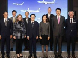 Eight professionals in formal attire posing on a stage in front of a blue screen with airplanes at a corporate event