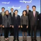 Eight professionals in formal attire posing on a stage in front of a blue screen with airplanes at a corporate event