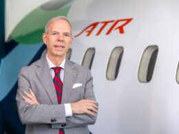 Executive in a gray suit and red striped tie stands with arms crossed beside an ATR aircraft in a hangar area