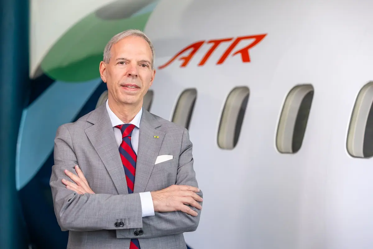 VLDB Executive in a gray suit and red striped tie stands with arms crossed beside an ATR aircraft in a hangar area