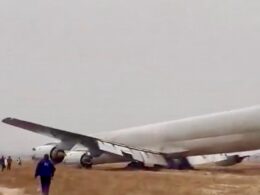 A large passenger jet lies wrecked on a dirt airfield wings damaged and fuselage crumpled while several people stand nearby observing