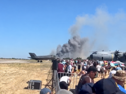 Crowd of spectators at an air show watches a smoky plume rise from near a parked US Air Force cargo plane beside a fighter jet on a sunny tarmac