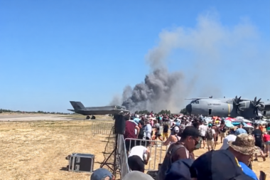 Crowd of spectators at an air show watches a smoky plume rise from near a parked US Air Force cargo plane beside a fighter jet on a sunny tarmac