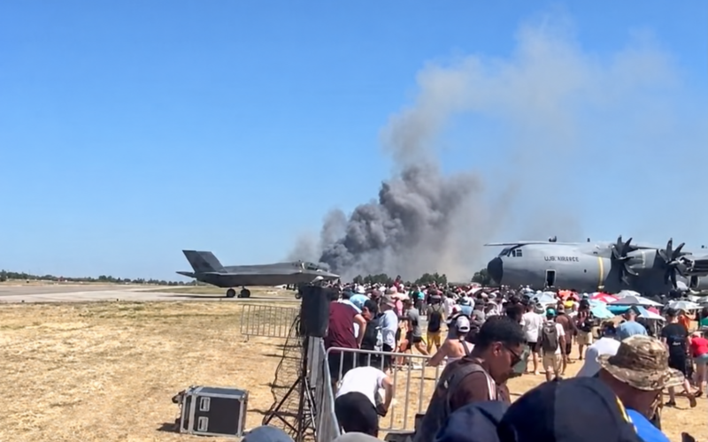 greece crash Crowd of spectators at an air show watches a smoky plume rise from near a parked US Air Force cargo plane beside a fighter jet on a sunny tarmac