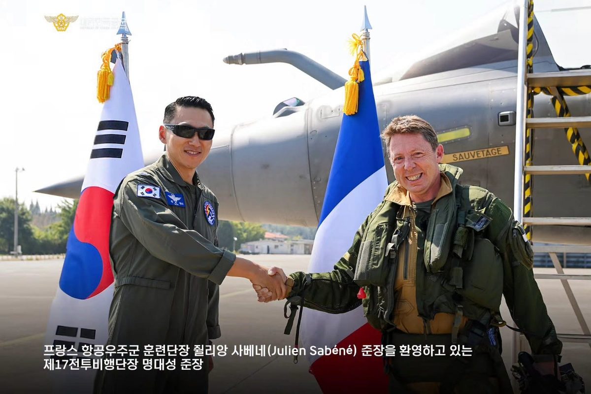 image Two pilots shake hands on a sunlit tarmac in front of a fighter jet with Korean and French flags nearby