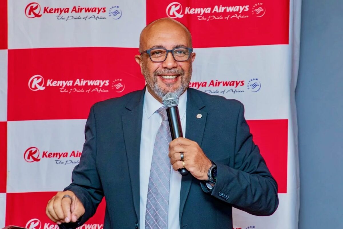 Man in a suit speaks into a microphone at a Kenya Airways event backdrop with red and white branding