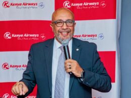 Man in a suit speaks into a microphone at a Kenya Airways event backdrop with red and white branding