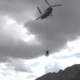 Military helicopter with long line lowering a suspended load over a rocky mountain landscape logo reading Ejército de Tierra in the corner