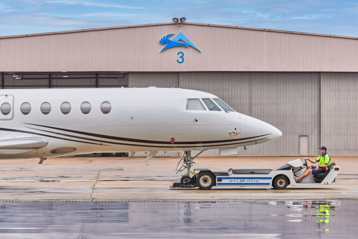 tv5511 0407 large67762ccf10f267b0b439ff000019a441 White private jet on the tarmac in front of a hangar with a blue logo and the number 3 a ground crew member in a neon vest sits on a tow tractor beside the nose