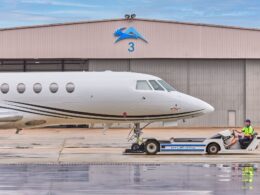 White private jet on the tarmac in front of a hangar with a blue logo and the number 3 a ground crew member in a neon vest sits on a tow tractor beside the nose