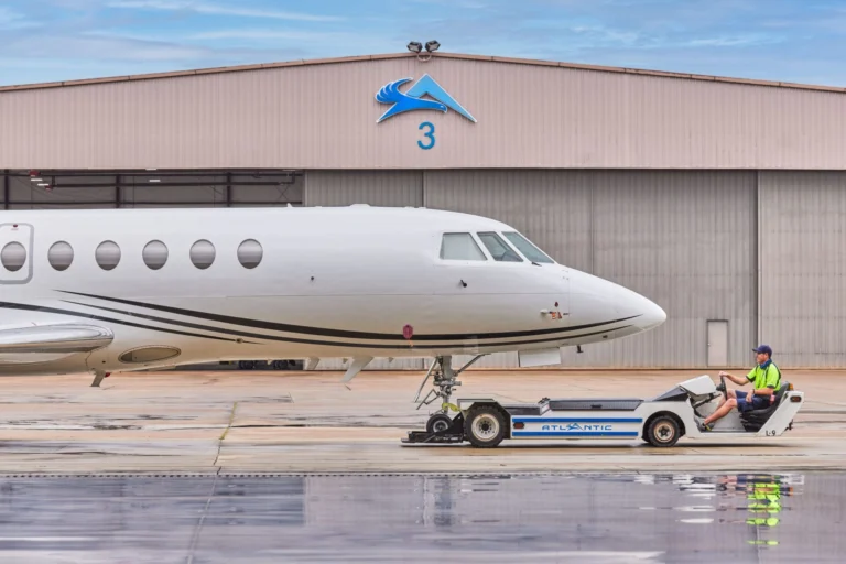 White private jet on the tarmac in front of a hangar with a blue logo and the number 3 a ground crew member in a neon vest sits on a tow tractor beside the nose