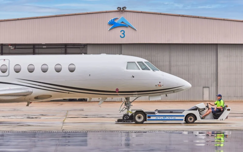 White private jet on the tarmac in front of a hangar with a blue logo and the number 3 a ground crew member in a neon vest sits on a tow tractor beside the nose