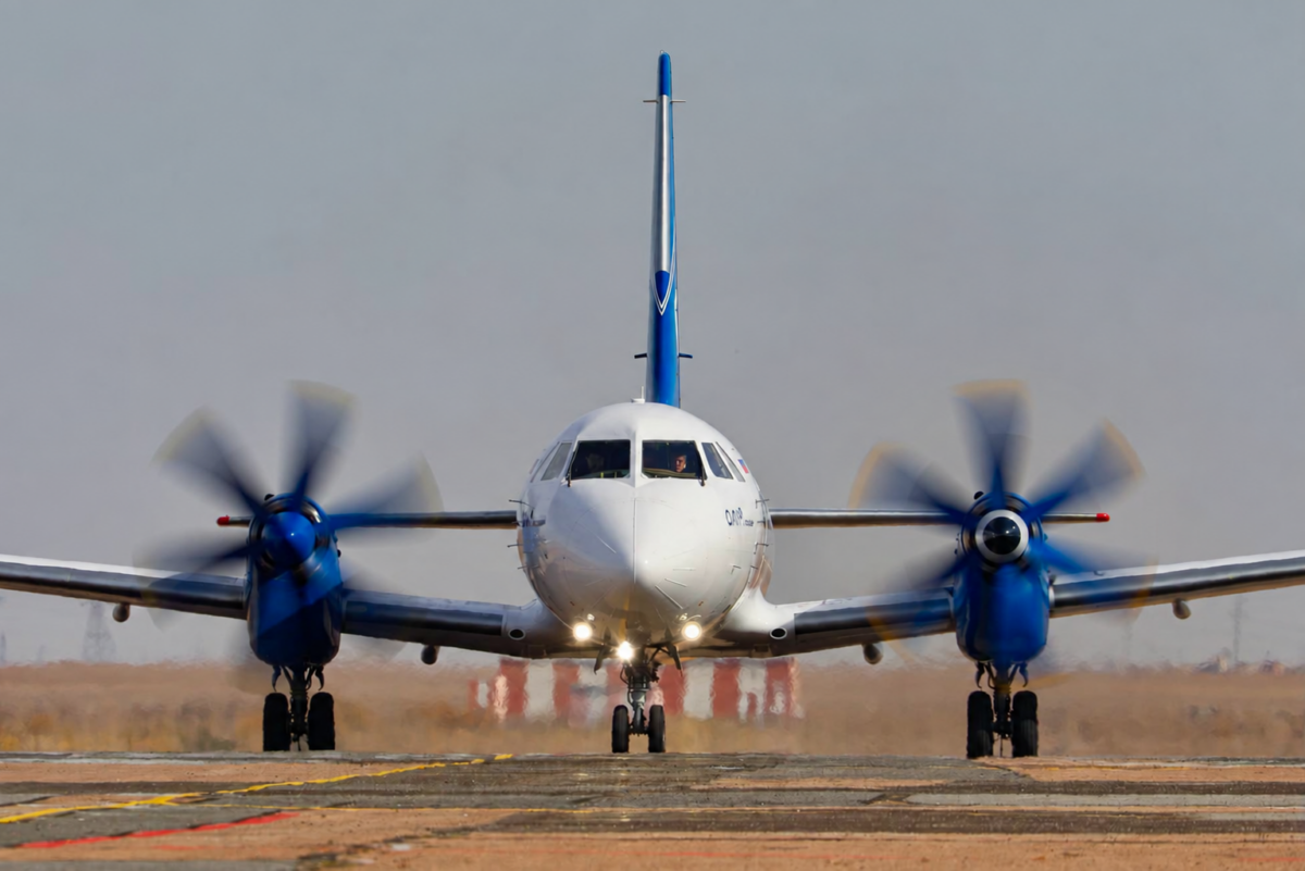 Front on view of a white twin engine turboprop aircraft with blue accents taxiing on a runway propellers spinning
