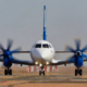 Front on view of a white twin engine turboprop aircraft with blue accents taxiing on a runway propellers spinning