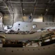 Maintenance hangar with a white aircraft fuselage labeled G EVTA undergoing assembly or repair surrounded by tools and equipment