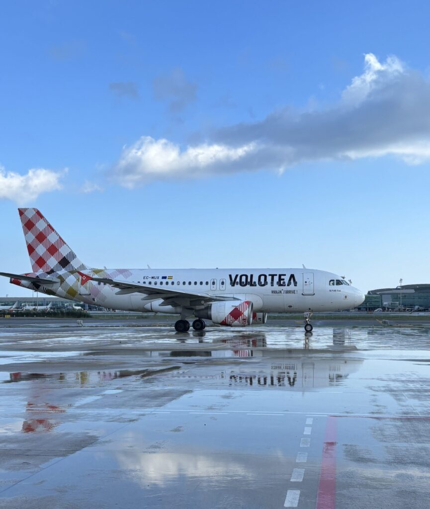Volotea airplane on a wet runway with a colorful patterned tail blue sky above and clouds nearby