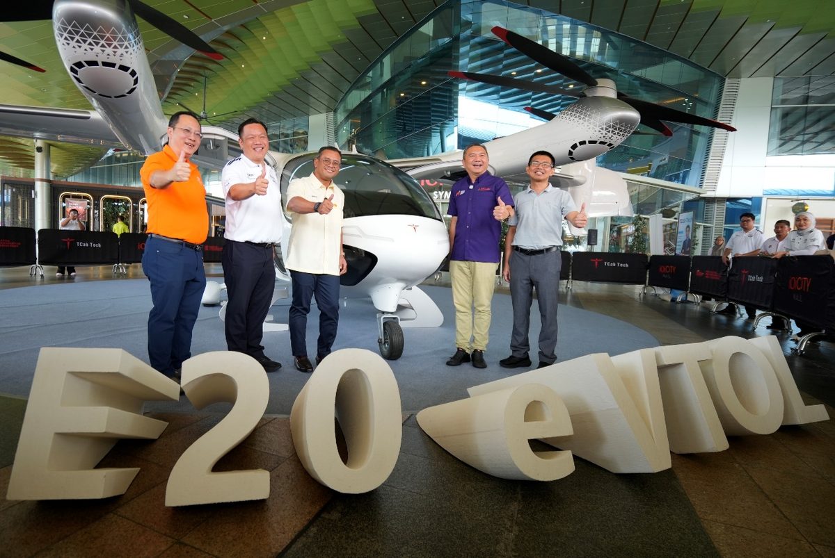 04 E20 eVTOL Group Photo Group of six men posing with thumbs up in an aviation showroom in front of a small white aircraft and large E 20 event letters on the floor