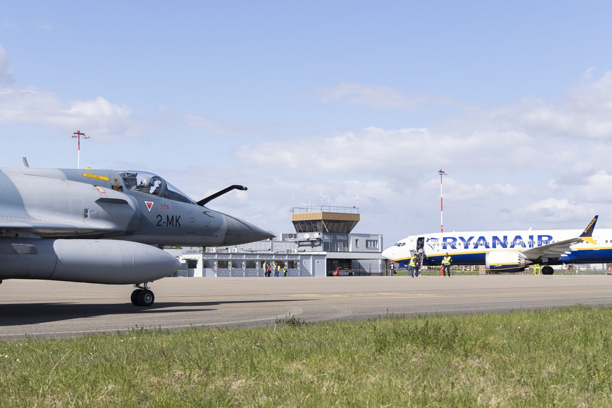 BASEX FRA ACE Gray military jet parked on a runway with its cockpit open a Ryanair airliner in the background and airport staff visible under a blue sky