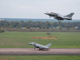 Military fighter jet lifting off from runway as another jet remains on the taxiway with a grassy field and tree line in the background