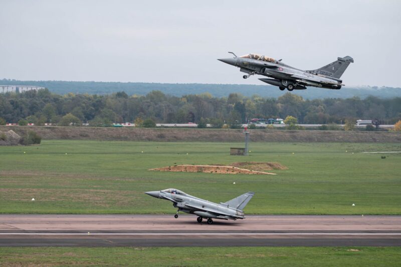 A French Air and Space Force Rafale B and a Royal Air Force Eurofighter Typhoon take off during a joint nuclear deterrence exercise in October 2026 Military fighter jet lifting off from runway as another jet remains on the taxiway with a grassy field and tree line in the background