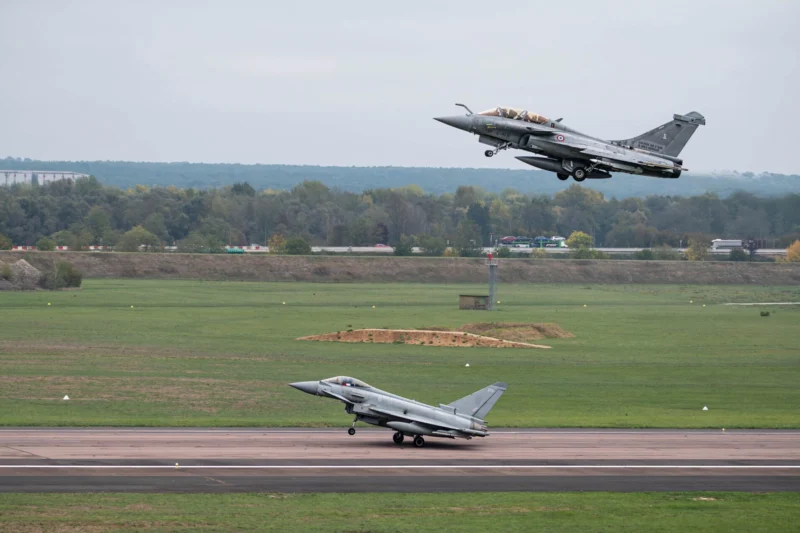 Military fighter jet lifting off from runway as another jet remains on the taxiway with a grassy field and tree line in the background