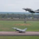 Military fighter jet lifting off from runway as another jet remains on the taxiway with a grassy field and tree line in the background