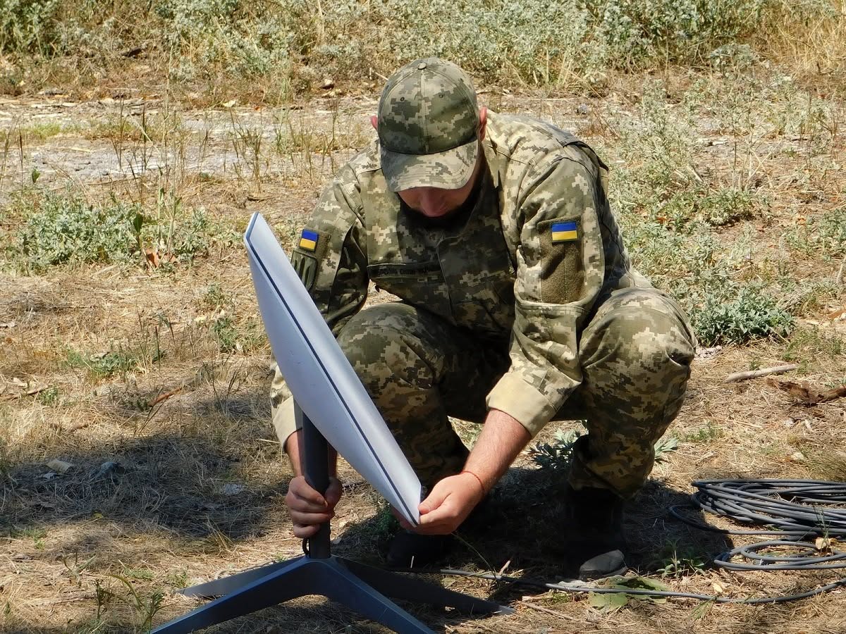 A Ukrainian serviceman installs a Starlink satellite communications terminal Soldier in camouflage kneels outdoors assembling a portable antenna or surveying device on a tripod