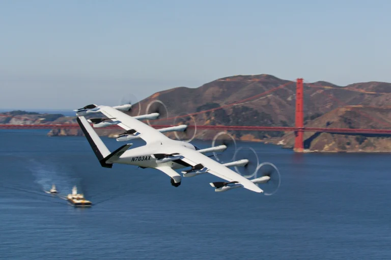 White tilt rotor aircraft flying low over the bay with a red suspension bridge and hills in the background