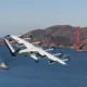 White tilt rotor aircraft flying low over the bay with a red suspension bridge and hills in the background