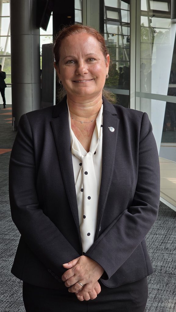Copy of 20260922 110758 Smiling woman in a black blazer and white polka dot blouse with a lapel pin standing in a glass walled lobby