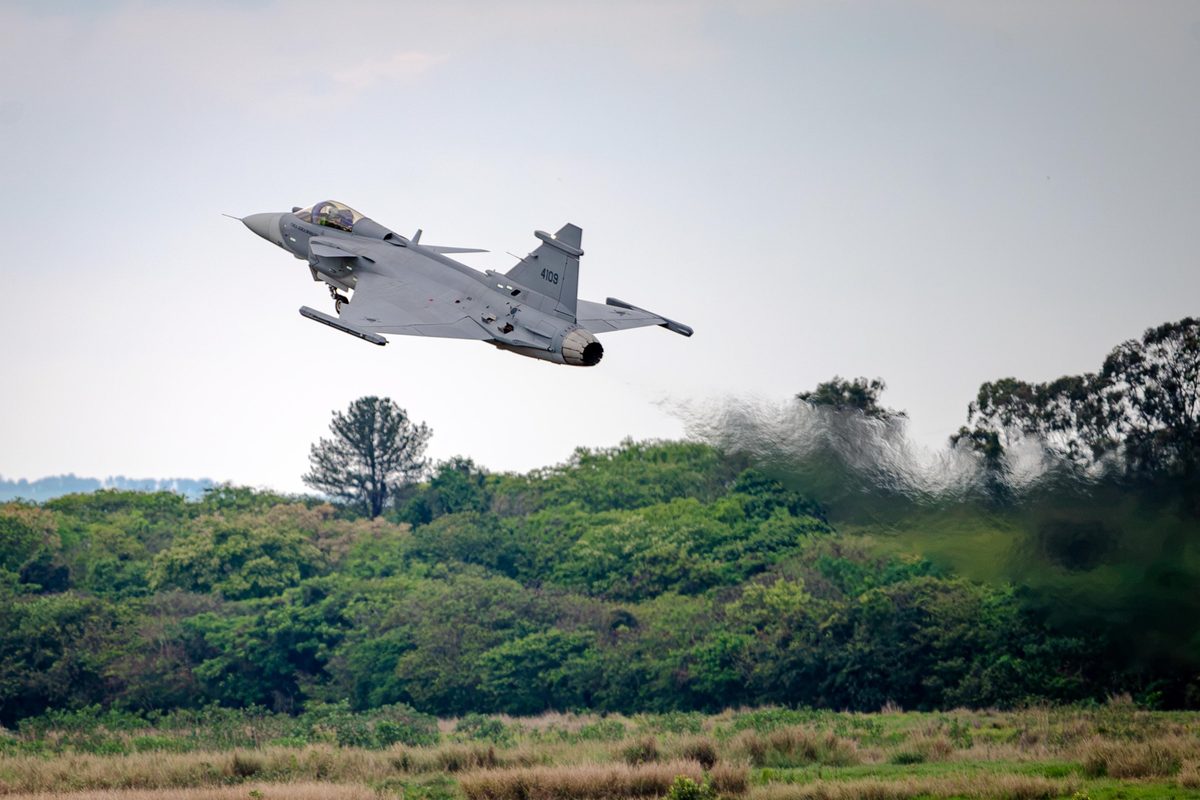First Gripen E produced in Brazil by Embraer makes its maiden flight 2 Gray military fighter jet lifting off above a forested field wings angled upward Tail number 4109 visible on the fuselage